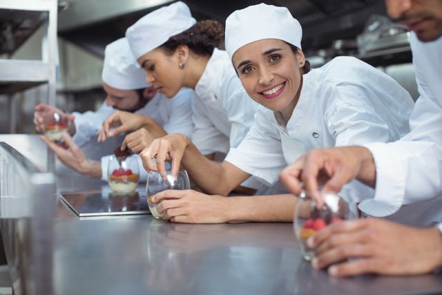 Chefs in a restaurant kitchen are finishing desserts in glass containers, showcasing teamwork and culinary skills. Ideal for use in articles about culinary arts, restaurant operations, teamwork in professional kitchens, or promotional materials for culinary schools and hospitality industry.