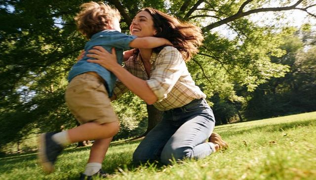 Mother embracing young son in sunny park kneeling on grass under large tree