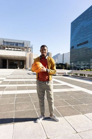 African american man holding basketball in plaza wearing headphones and mustard jacket