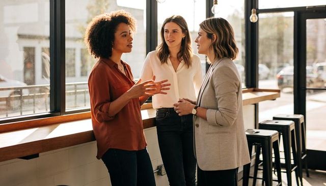 Three women collaborating in casual coworking space by sunlit windows during business conversation
