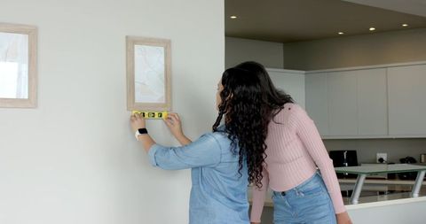 Diverse female duo aligning wall art with spirit level at home
