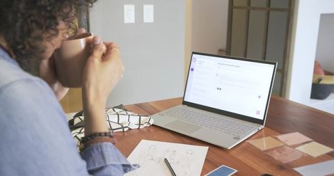 Non-binary designer working from home holding mug at laptop with sketches and swatches