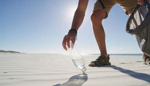 Volunteer picking up plastic bottle on sandy beach during coastal cleanup, backlit sun