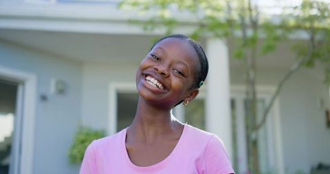 Smiling teen girl tilting head on suburban porch in pink tee, joyful lifestyle portrait