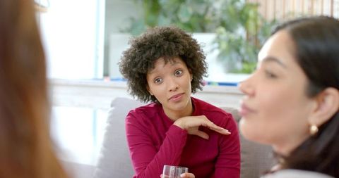 Diverse women chatting on sofa at home, candid conversation in sunlit lounge