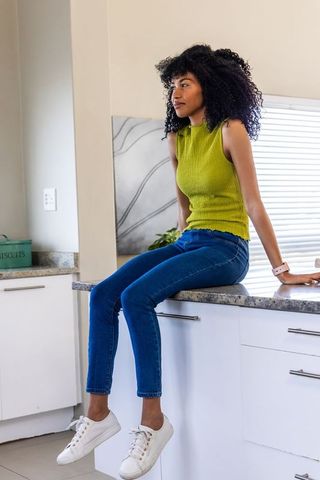 Curly haired woman sitting on kitchen counter wearing green top and jeans, looking right