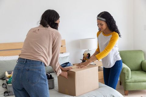 Mother and teenage daughter packing and sealing moving box in bright bedroom