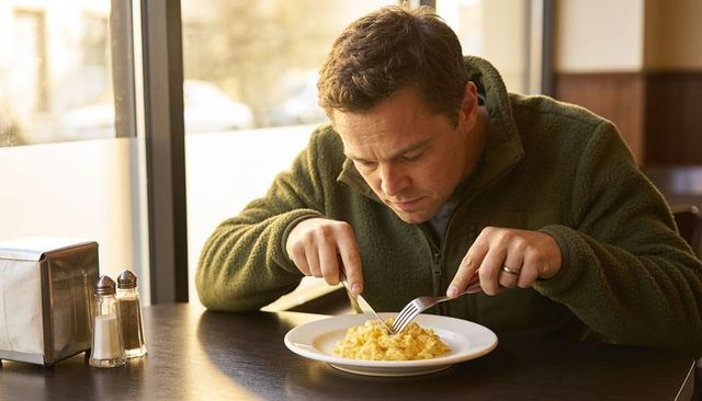 Man cutting scrambled eggs at sunlit diner window, cozy solo breakfast in cafe booth