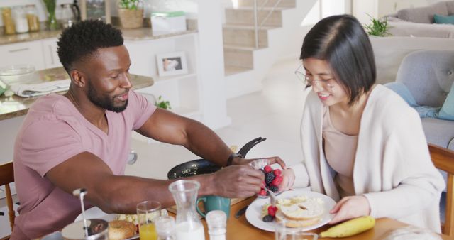Couple smiling at each other while enjoying breakfast in a cozy home setting. They are eating homemade food and sharing a joyful moment, highlighting a happy and healthy relationship. Ideal for use in contexts such as healthy eating campaigns, relationship counseling, lifestyle blogs, or marketing materials focused on domestic life and love.