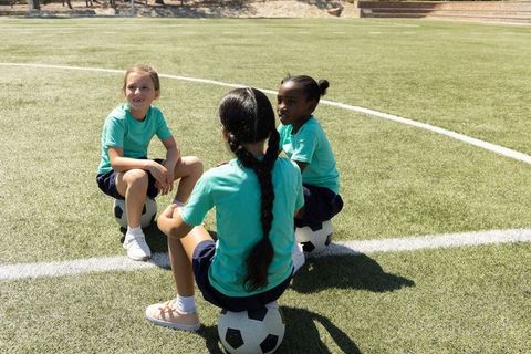 Young girls sitting on soccer balls chatting on turf during sunny soccer practice