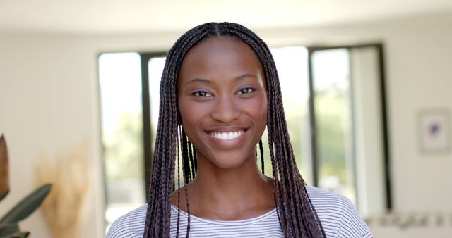 Young woman with braided hair standing in a sunlit room, smiling warmly. Perfect for lifestyle blogs, self-confidence articles, promotional material, mental health campaigns, and any content focusing on happiness or positivity.