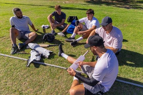 Multicultural cricket team sitting on grass inspecting bat and pads, discussing tactics