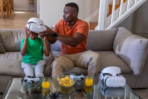 African American Father Helping Child Wear VR Headset on Sofa with Controllers and Snacks
