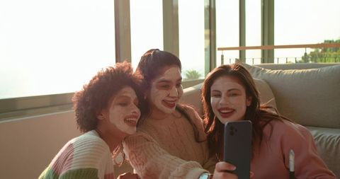 Diverse friends wearing face masks and knitwear taking cozy sunlit selfie on sofa