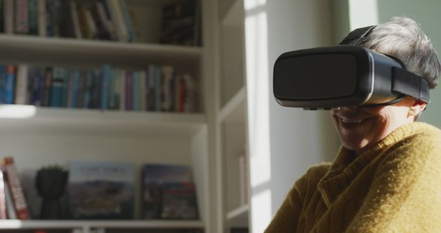 Elderly woman smiling while using a VR headset in the comfort of her home. Bookshelves filled with books in the background. Ideal for use in technology, home comfort, or senior lifestyle themes.