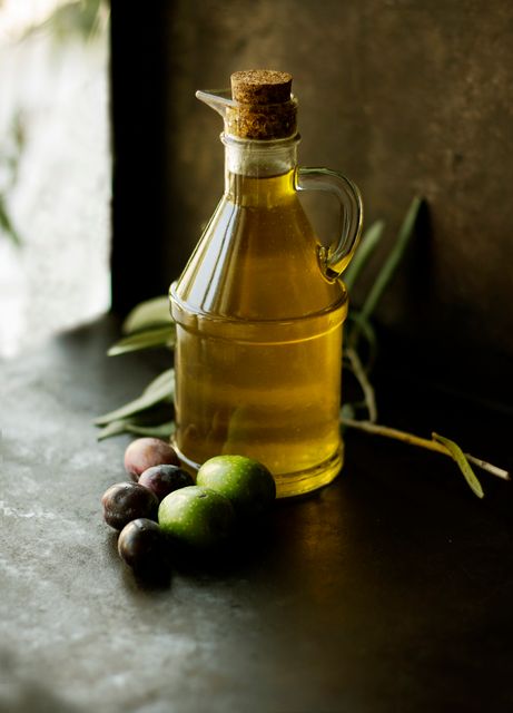 Rustic scene features glass bottle filled with olive oil accompanied by fresh green and black olives and olive leaves. Ideal for use in cooking blogs, recipes, Mediterranean cuisine promotions, and healthy eating campaigns. Emphasizes natural and wholesome elements suitable for gourmet and organic product advertisements.