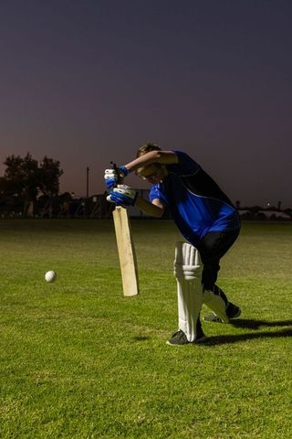 Teen cricketer blocking low ball at twilight field with pads and gloves practicing defense