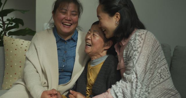 Three Asian women—an elderly woman, a middle-aged woman, and a young adult—are sitting on a sofa, smiling and laughing together, conveying a joyful and affectionate family bonding moment. Perfect for illustrating themes of family, togetherness, happiness, and multigenerational relationships. Use this for advertisements, blog posts, or social media content that focus on family life, support, and joy in community.