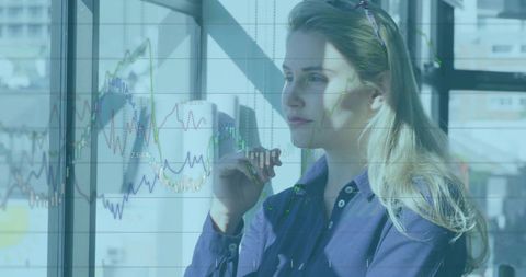 Gazing businesswoman holding pen while analyzing financial charts through office window