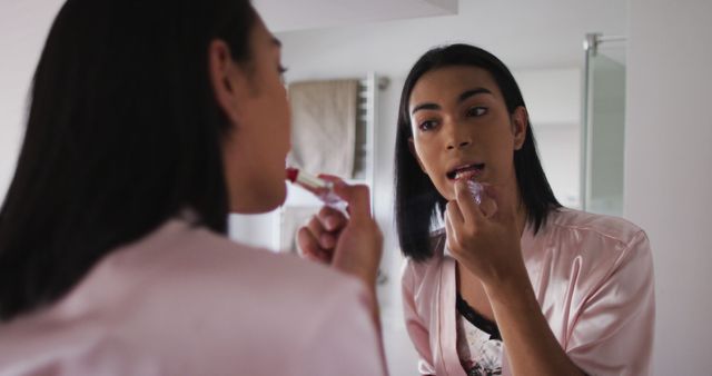 Woman stands in front of bathroom mirror applying lipstick, seemingly engaged in morning beauty routine. She is wearing a silk robe, indicating a serene and self-care focused moment. Useful for illustrating themes around beauty routines, self-confidence, personal care, and cosmetics advertisements.