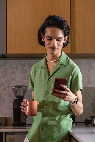 Asian man in green shirt holding mug and smartphone checking messages in modern kitchen
