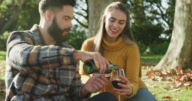 Couple sitting on grass enjoying a bottle of wine in an autumn park. Man wearing plaid shirt pouring wine into glass held by woman in mustard-yellow sweater. Perfect for promoting picnic-related products, autumn outings, couples' activities, or advertising leisure lifestyle and relaxation in natural settings.