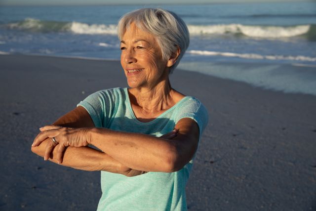 Senior Caucasian woman enjoying a sunny day at the beach, stretching and exercising with the sea in the background. Ideal for promoting healthy lifestyles, fitness for seniors, beach vacations, and wellness activities.