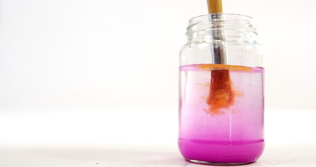 This image shows a paintbrush submerged in a water jar with purple paint dissolving in water. The scene represents the cleaning process of an artist while creating artwork. It is perfect for art-related content, tutorials, or illustrating steps in a painting process. Great for educational materials, blogs, or articles focused on painting techniques and tools.