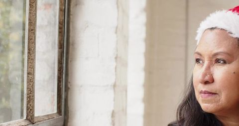 Mature woman wearing santa hat gazing through aged metal window frame during christmas