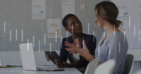 Two female professionals discussing strategy in boardroom using tablet and laptop