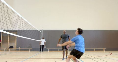 Male volleyball players competing at net in gymnasium, blue-shirt preparing to receive ball