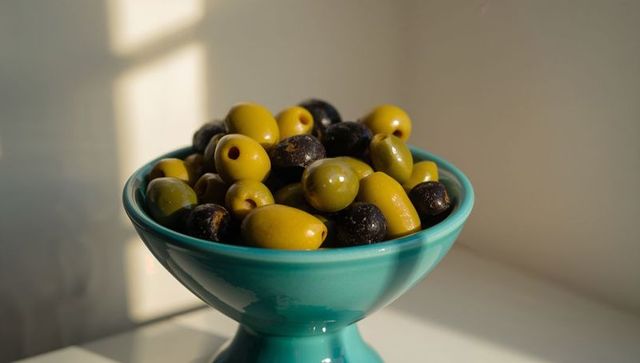 Sunlit turquoise bowl overflowing with glossy pitted green and black olives, closeup