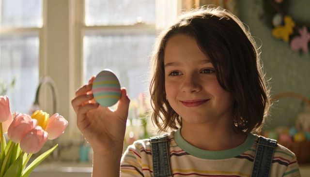 Smiling child holding pastel-striped easter egg by sunlit kitchen window with tulips, morning light