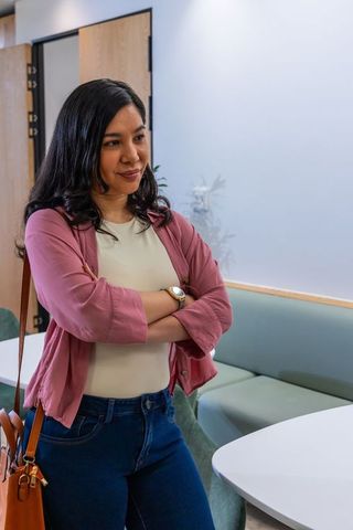 Woman standing with arms crossed in modern office lounge carrying tan shoulder bag