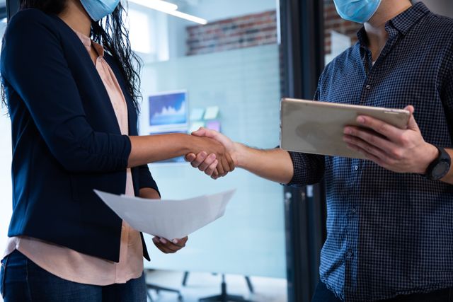 Colleagues in a casual office environment are shaking hands while wearing face masks, emphasizing safety and health protocols during the Covid-19 pandemic. This image can be used to illustrate workplace safety measures, business collaboration, and teamwork during the pandemic. It is suitable for articles, blogs, and presentations related to office work, pandemic precautions, and professional interactions.