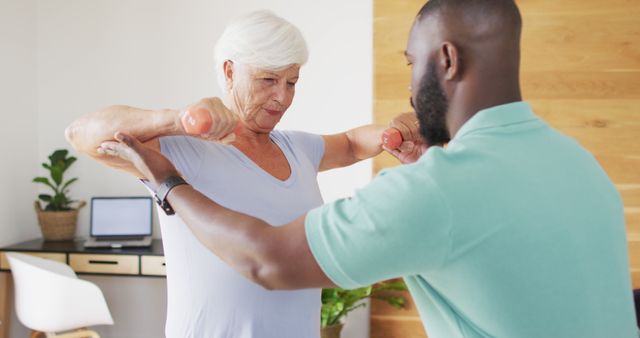 Senior woman exercising with personal trainer using dumbbells indoors, promoting strength training and elderly fitness. Useful for websites focused on fitness, aging, health, personal training, and wellness programs.