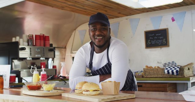 Chef wearing striped apron stands at counter in food truck, presenting freshly prepared burgers with pride. Great for use in articles about street food entrepreneurship, small business promotion, outdoor food events, or cooking showcases.