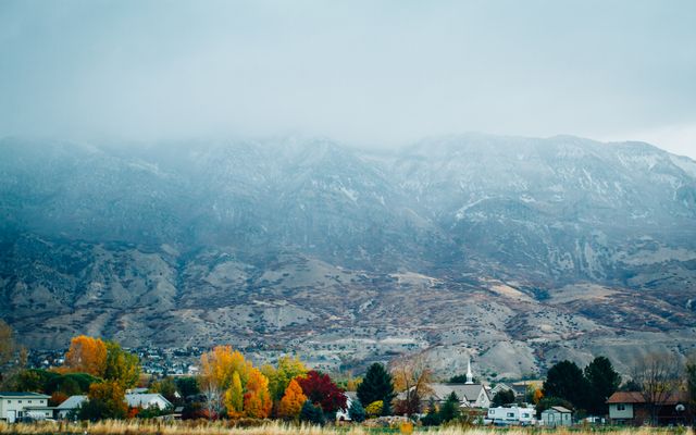 Perfect for illustrating small-town life, seasonal changes, or the beauty of rural landscapes, this image highlights a charming town nestled in mid-autumn near a mountain range partially obscured by clouds. The mix of vibrant fall colors against the mountainous backdrop makes it suitable for travel blogs, seasonal promotions, or nature-related content.