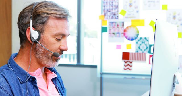 This image shows a mature male customer support agent wearing a headset, typing on a computer in office setting. It is useful for illustrating concepts related to customer service, call center operations, and helpdesk support. Ideal for business training materials, service-oriented websites, and professional publications.