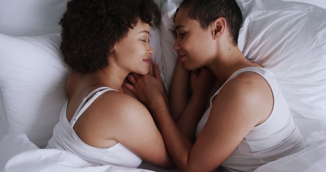 Two women are lying in bed facing each other, holding hands and smiling. Both are wearing white tops and look content. This image can be used for content relating to relationships, love, affection, LGBT+ couples, and intimate moments.