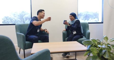 Diverse healthcare colleagues clinking coffee cups in relaxed break lounge wearing scrubs