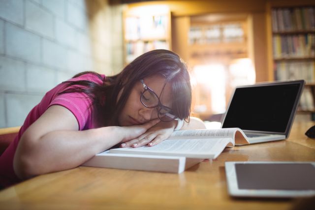 Young female student taking a nap on an open book at a desk in a library. She appears tired and exhausted from studying, with a laptop and tablet nearby. This image can be used to depict themes of education, student life, academic stress, and the importance of rest. Ideal for articles, blogs, and advertisements related to student well-being, study tips, and academic resources.