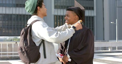 Graduate celebrating diploma on rooftop while friend adjusting mortarboard