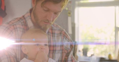 Cradling bearded father gazing at infant with sun flare in warm backlit kitchen