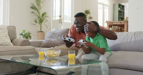 Father and son playing video games on sofa with snacks and orange juice, bonding