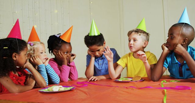 Children sitting at a table during a birthday party, wearing colorful hats and talking. The diverse group of friends appears to be enjoying their time together. An ideal image for themes related to childhood celebrations, friendships, events, and happy moments.