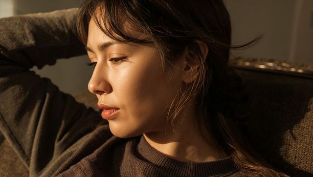 Sunlit profile of thoughtful asian woman resting in brown knit sweater, warm golden light