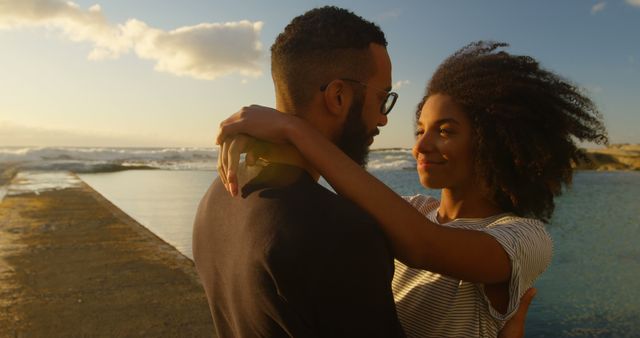 Romantic couple embracing on shore while enjoying sunset. Useful for love, romance, travel, or lifestyle themes in promotions and advertisements depicting intimate moments, peaceful interactions, and serene environments.