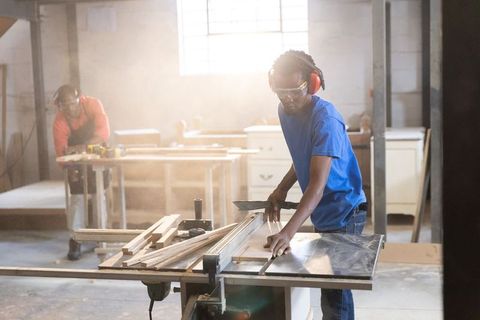 African american men cutting wood on table saw in carpentry workshop wearing safety gear