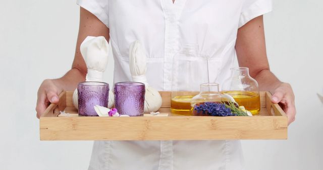 A Caucasian woman in a white uniform presents a wooden tray with spa essentials, including candles, oils, and lavender, with copy space. Her attire and the items suggest she may be a spa therapist ready to provide a relaxing experience.